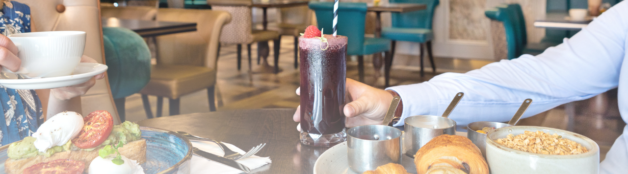 Hand holding a fruit juice with a plate of pastries in front. To the left is mexican eggs with a hand holding a coffee